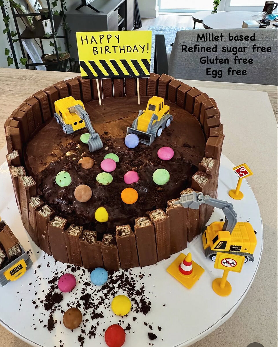 A digger-themed birthday cake with chocolate fingers, sweets and toy diggers, labelled millet based, refined sugar free, gluten free and egg free.