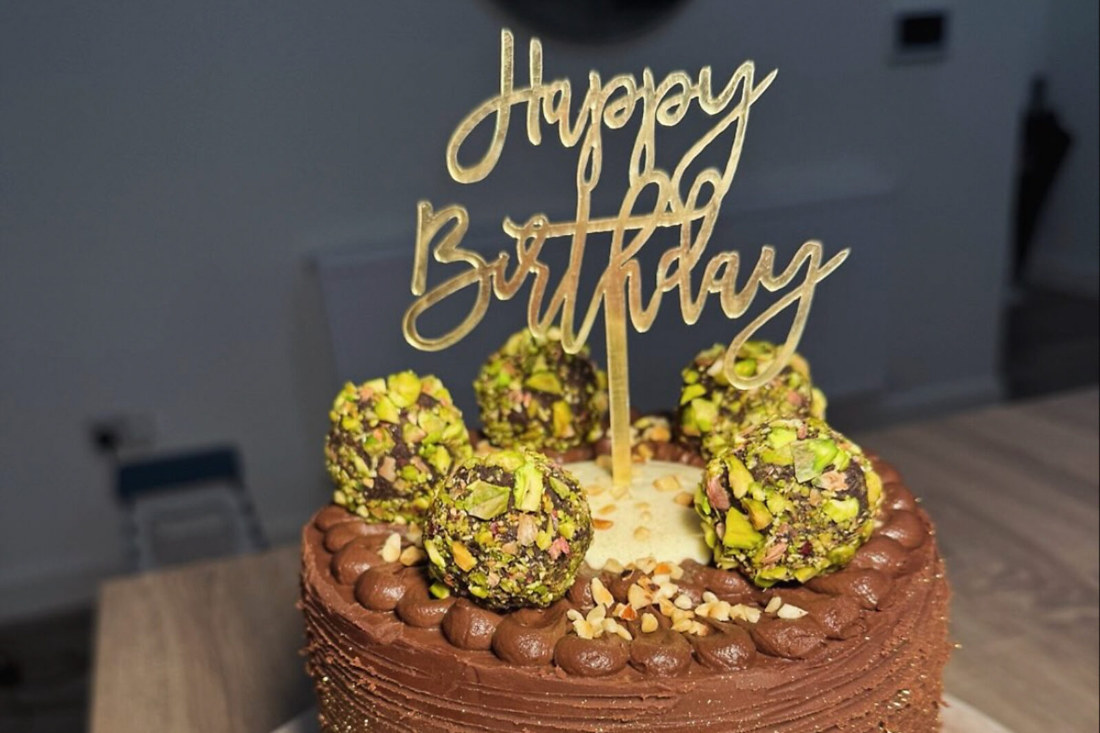 A tall chocolate ganache cake topped with pistachio-crusted truffles and a gold Happy Birthday topper.