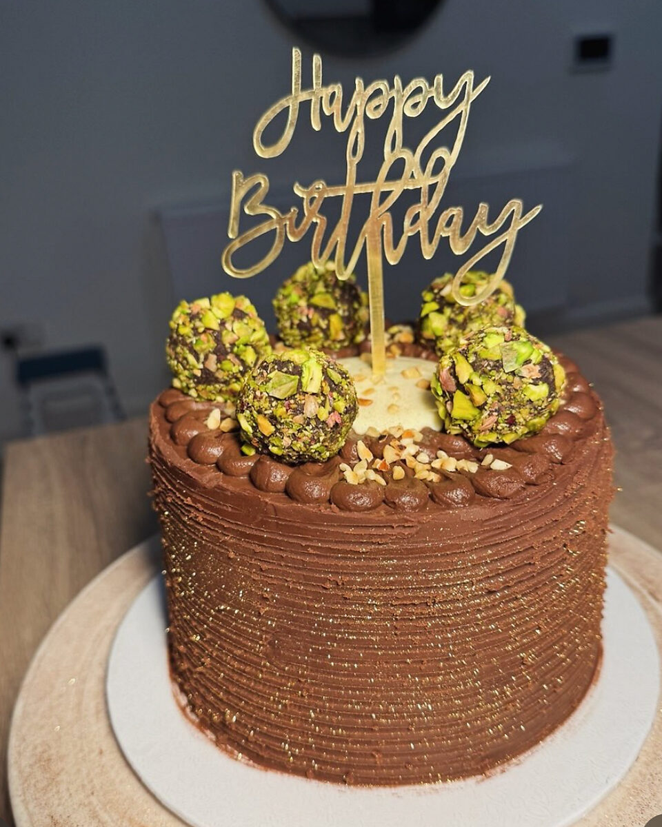 A chocolate cake with pistachio truffles and a gold Happy Birthday topper on a wooden table.